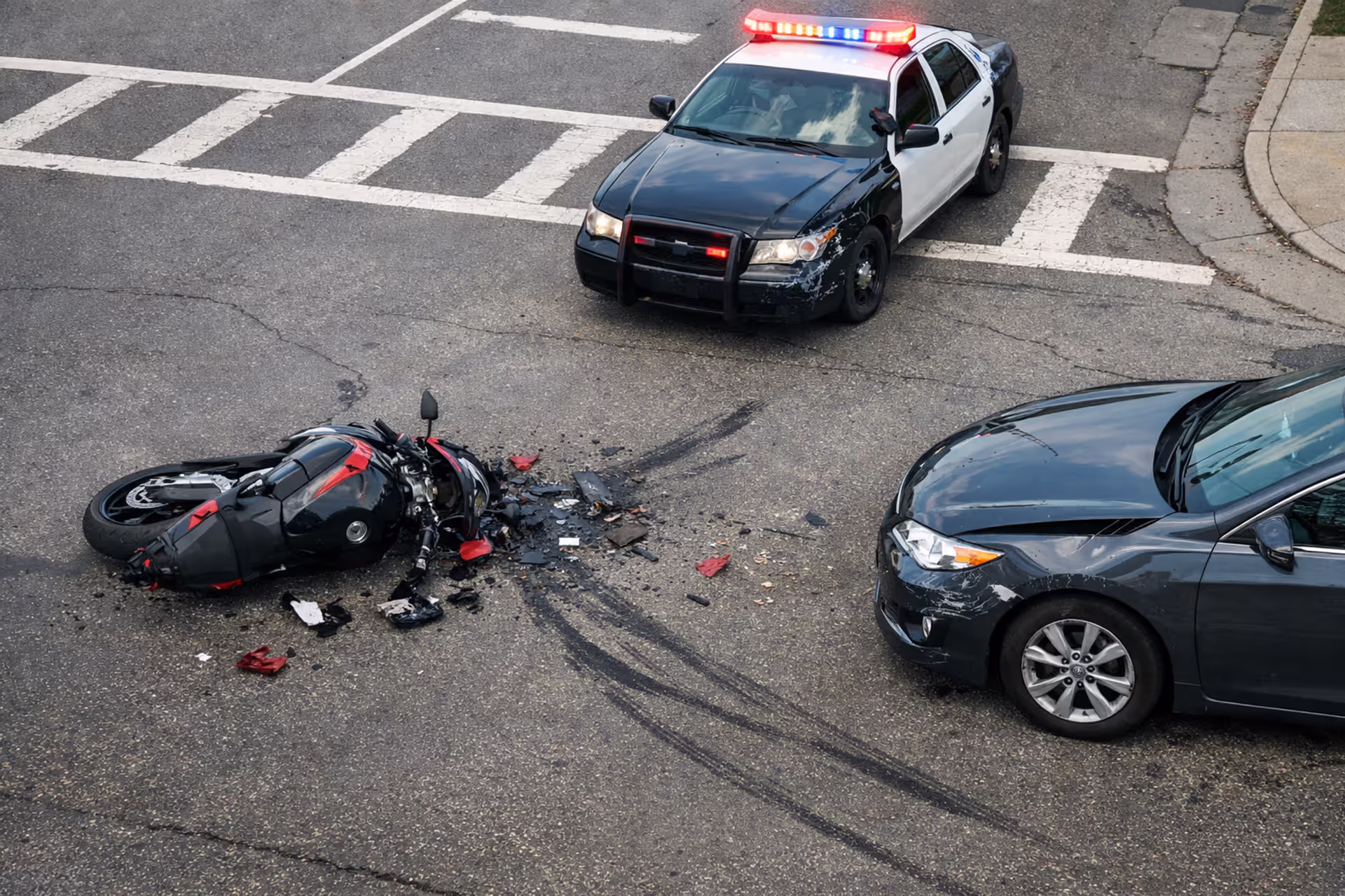 Damaged sport motorcycle lying on asphalt at urban intersection after collision with car, police vehicle with flashing lights in background